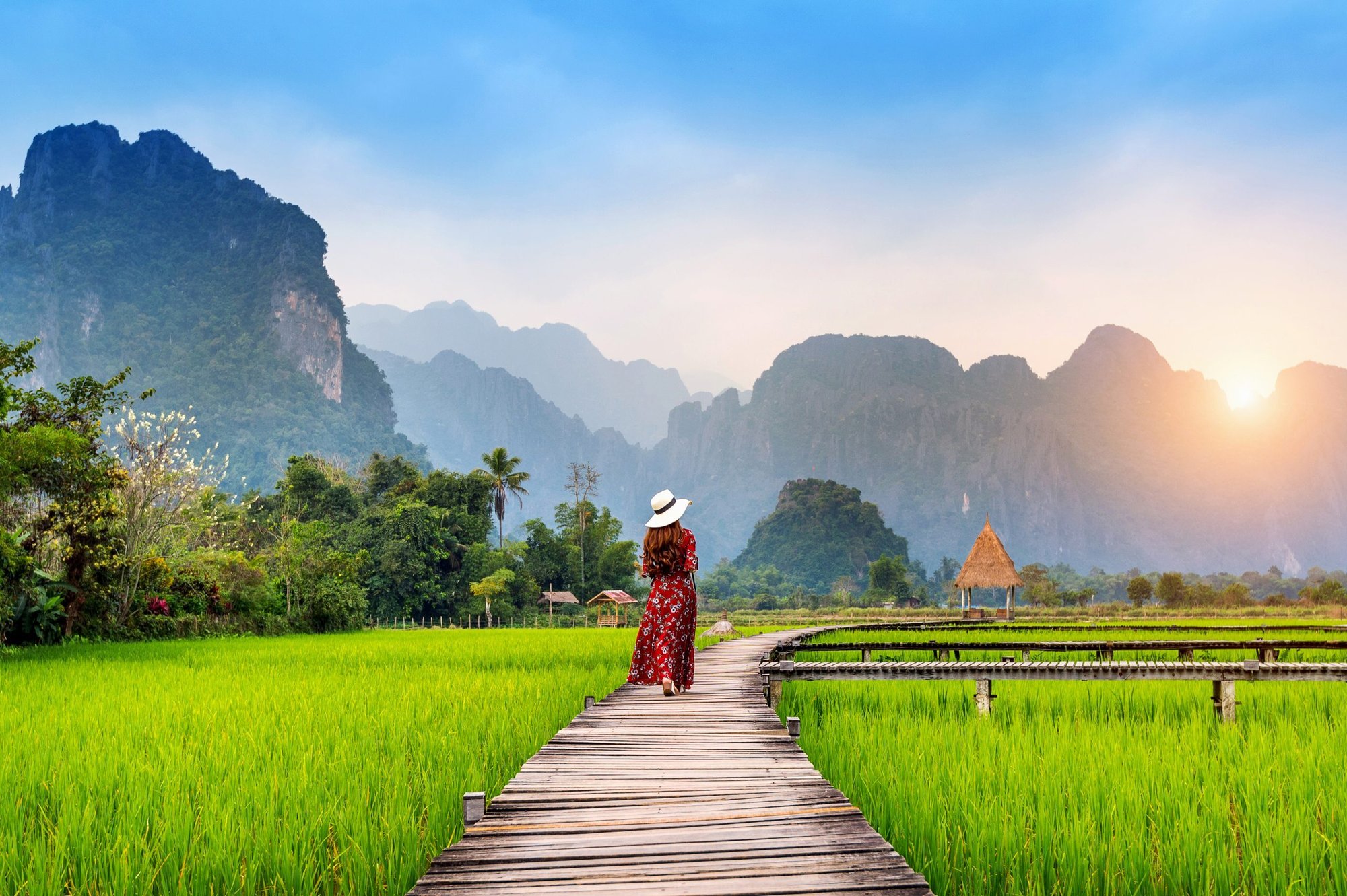 young-woman-walking-wooden-path-with-green-rice-field-vang-vieng-laos-scaled